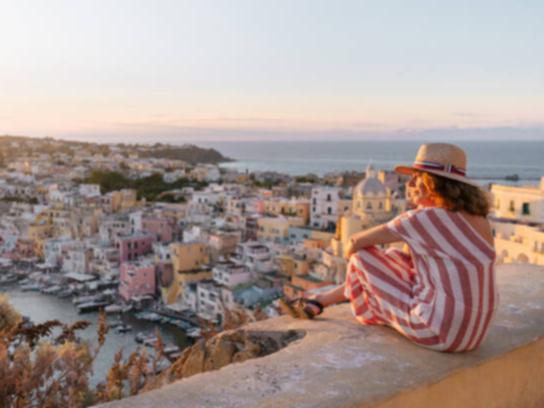 Side view of positive mature female traveler in striped dress and hat enjoying amazing seascape during sunset in Procida island.