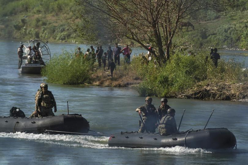 Migrants stand on an island in the middle of the Rio Grande as law enforcement personnel address them from a raft.