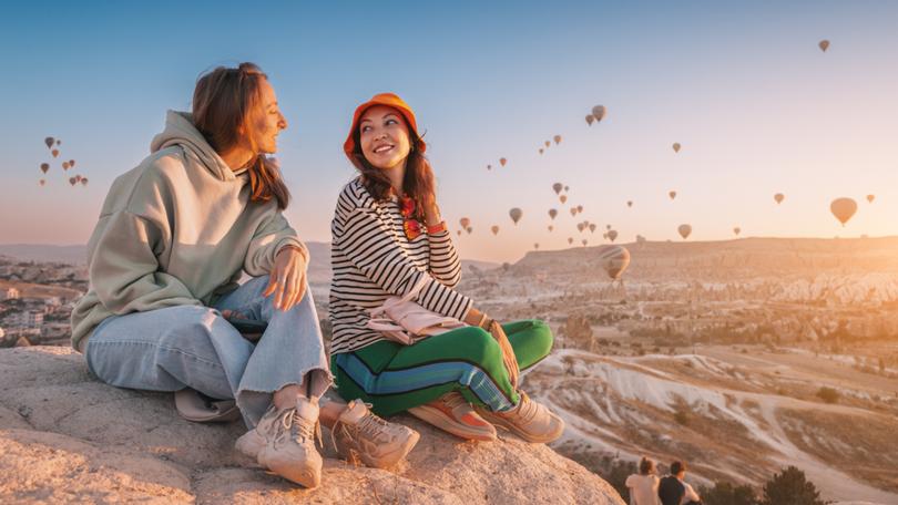 Girl friends sitting on a clifftop viewpoint and admiring view of majestic flying hot air balloons in Cappadocia