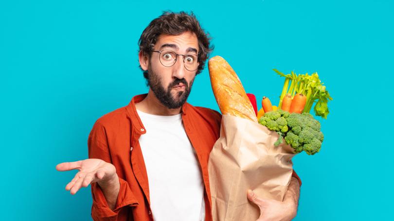 young bearded man feeling puzzled and confused, doubting, weighting or choosing different options with funny expression and holding a vegetables bag 