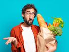 young bearded man feeling puzzled and confused, doubting, weighting or choosing different options with funny expression and holding a vegetables bag 