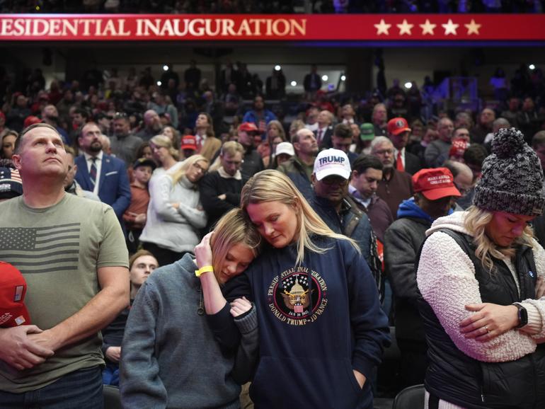 Supporters of President Donald Trump bow their heads during the invocation while watching inauguration ceremonies taking place at the U.S. Capitol on screens at Capitol One Arena in Washington, Monday, Jan. 20, 2025. (AP Photo/Mark Schiefelbein)