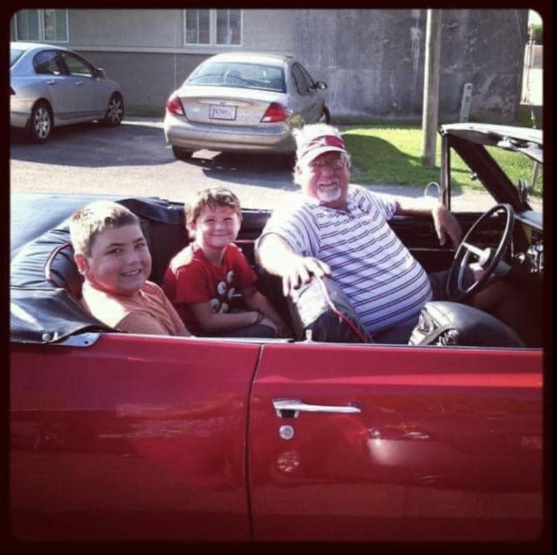 Bill Bryan driving a red convertible in 2018 with his grandsons in the back seat. 