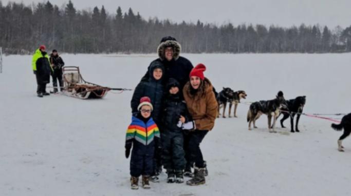 Patrice Poltzer poses with her husband and three boys for a family photo in Finland with a sled and dogs in the snowy backdrop.