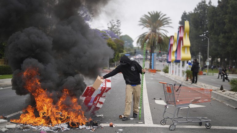 A protester places debris in a fire as Border Patrol personnel in riot gear and gas masks stand guard outside an industrial park in the city of Paramount, California.