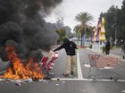 A protester places debris in a fire as Border Patrol personnel in riot gear and gas masks stand guard outside an industrial park in the city of Paramount, Calif., on Saturday, June 7, 2025 (AP Photo/Eric Thayer)