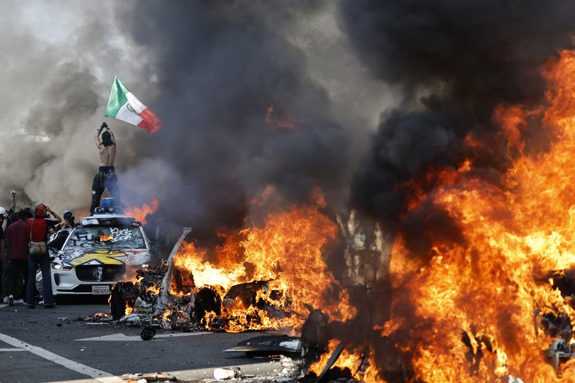 A protester holds up a Mexican flag as burning cars line the street on June 08, 2025 in Los Angeles, California.