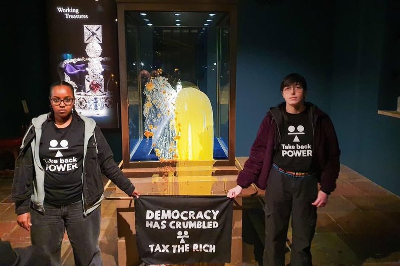 The pair stood in front of the defaced display holding a banner reading “Democracy has crumbled – Tax The Rich.”