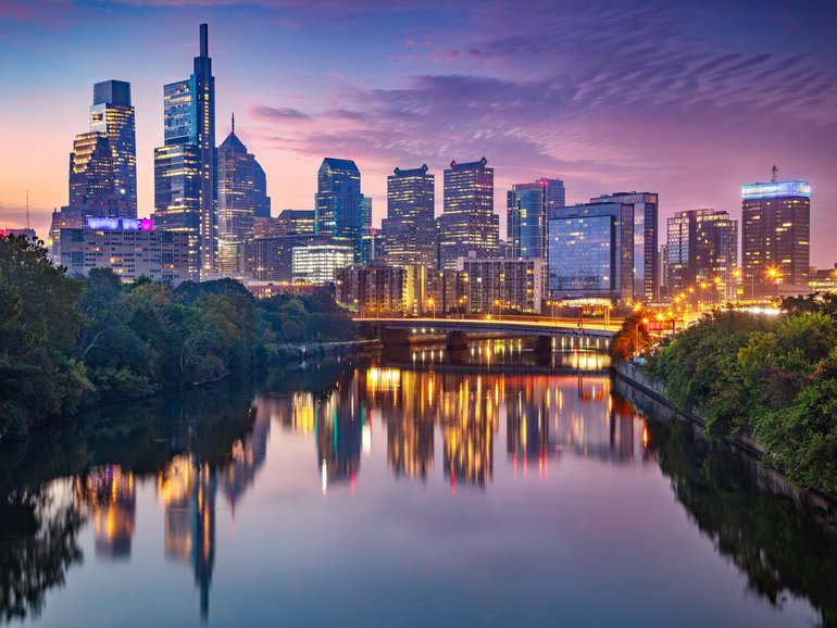 Philadelphia, Pennsylvania, USA at twilight. The city skyline is illuminated, with modern skyscrapers reflecting vibrant lights in the calm waters of the Schuylkill River. 