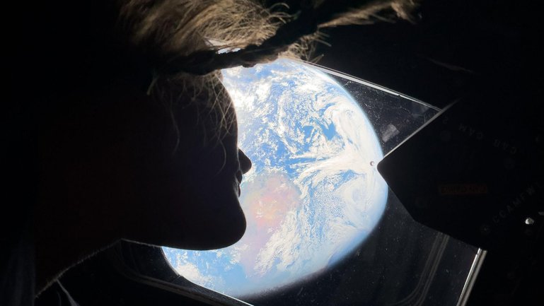 A photo provided by NASA shows NASA astronaut and Artemis II mission specialist Christina Koch as she peers out of one of the Orion spacecraft's main cabin windows, looking back at Earth, as the crew travels towards the Moon, April 4, 2026. The astronauts said they had lost track of which day it is on Earth on their transit to the moon. (NASA via The New York Times) — NO SALES; EDITORIAL USE ONLY—