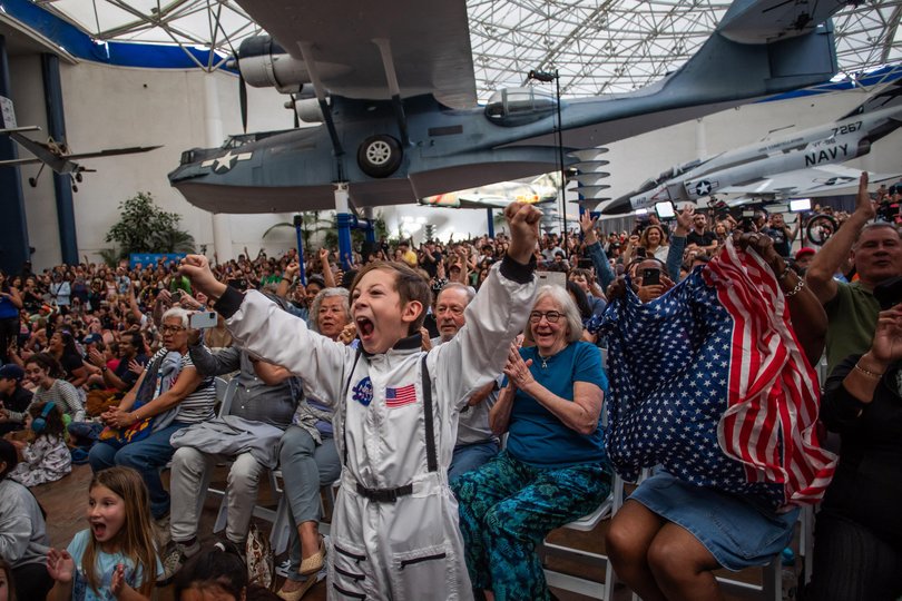 A young boy wearing an astronaut costume cheers next to a woman waving a flag as they watch a live broadcast of the return of the Artemis II crew members to Earth at the San Diego Air and Space Museum during a watch party for the crew's splash down in the Pacific Ocean, in San Diego, California, on April 10, 2026. 