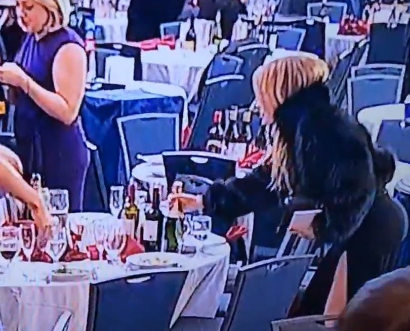 A well dressed blonde woman appears to steal a bottle of wine from an abandoned table