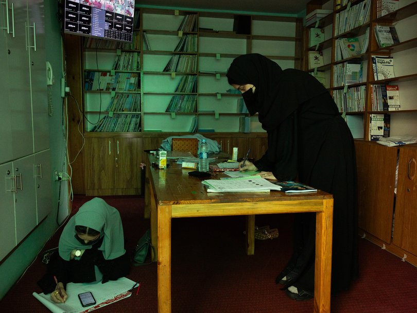 Teachers fill out administrative paperwork, at a school where a CCTV screen monitors classrooms.